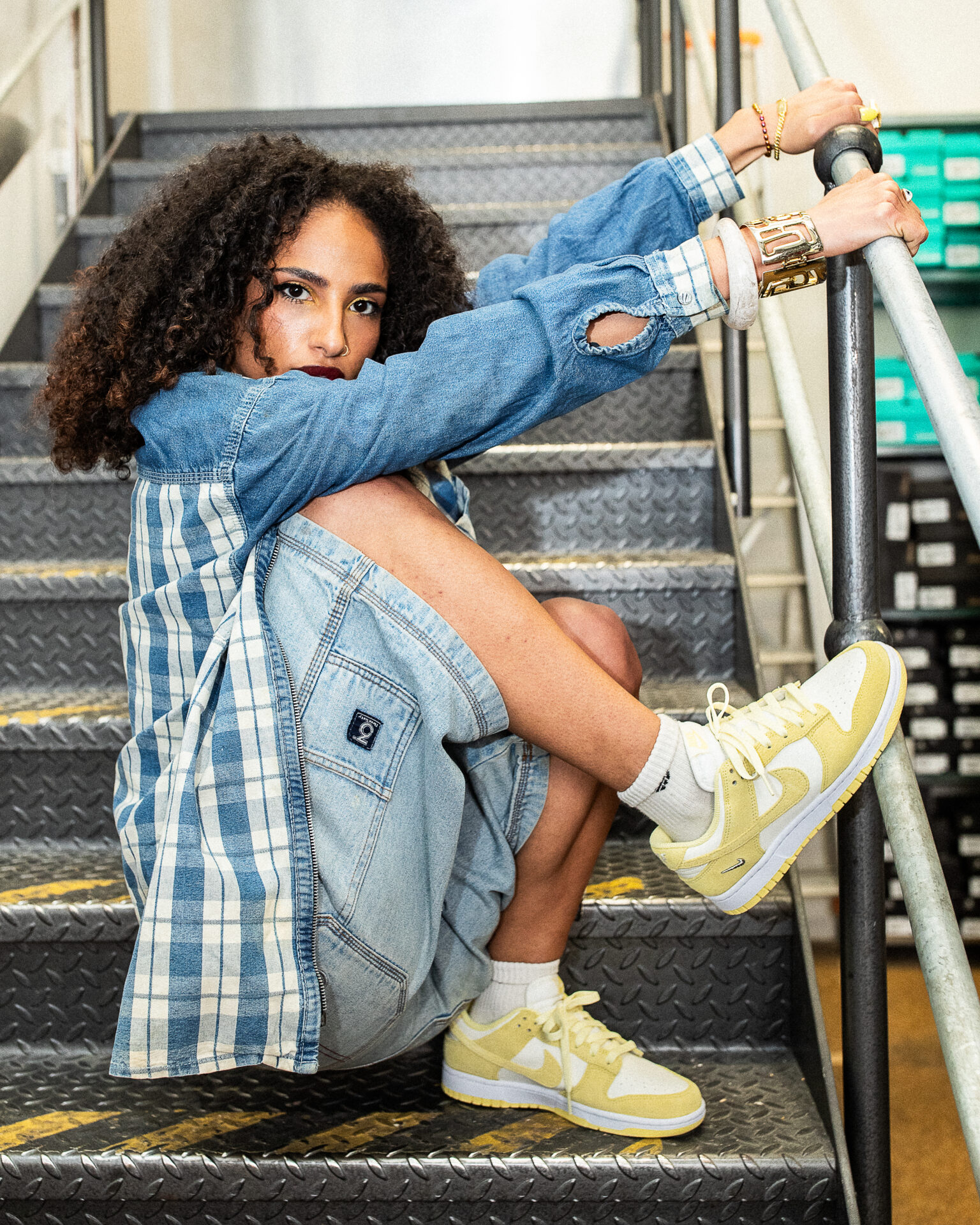 Roukaya sitting on stairs wearing jorts, a denim checked jacket, and white and yellow Nike trainers.