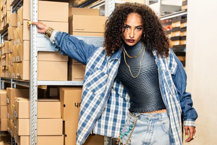 One of our creatives taking part in our Manchester window project, Roukaya B, standing in a stockroom full of schuh shoe boxes. She wears a textured blue top with a checked denim jacket and denim jorts, paired with gold jewellery.