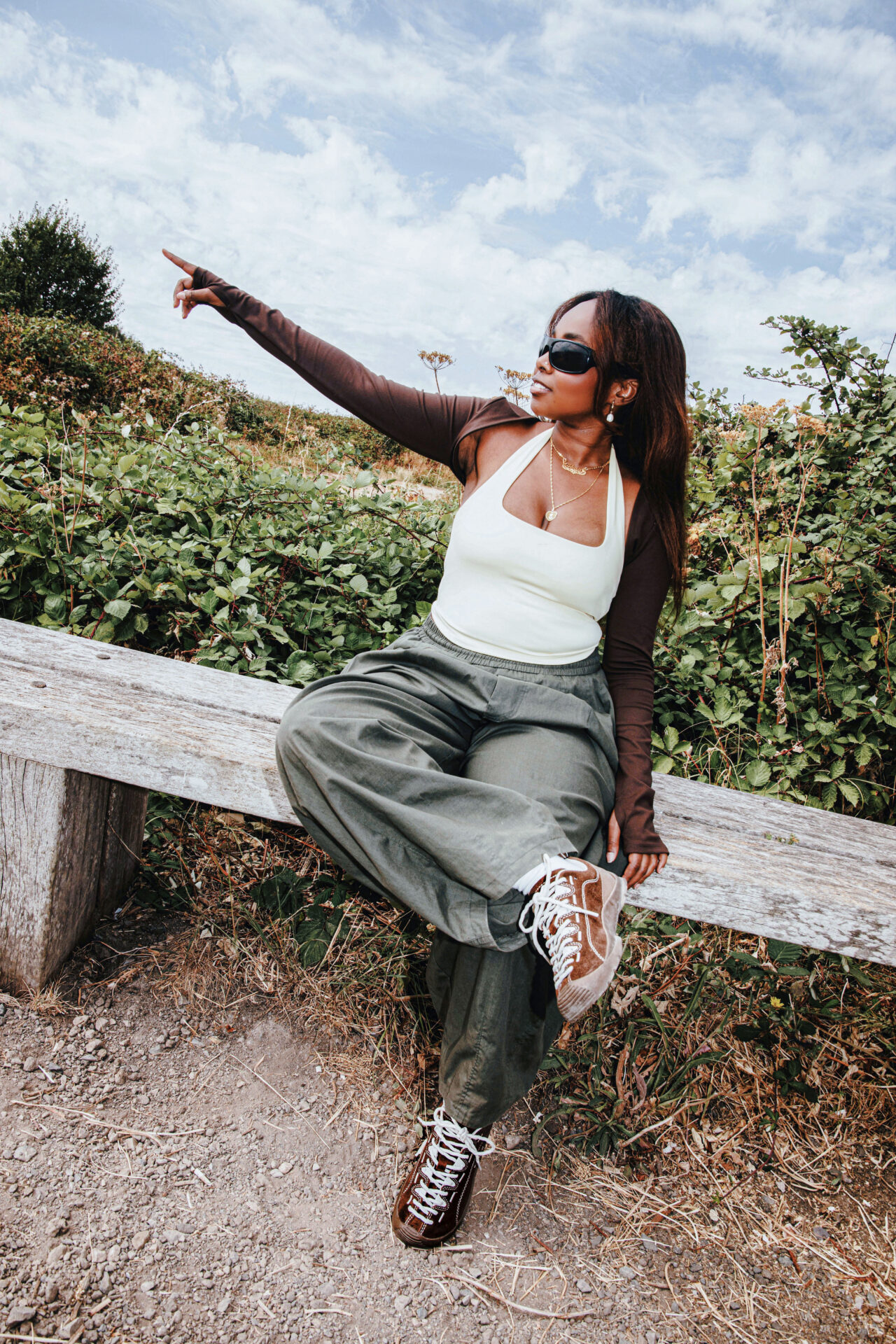 Person wearing khaki parachute pants, a white tank top, brown bolero style top, brown KEEN Jaspers, and black sunglasses, sitting outside.