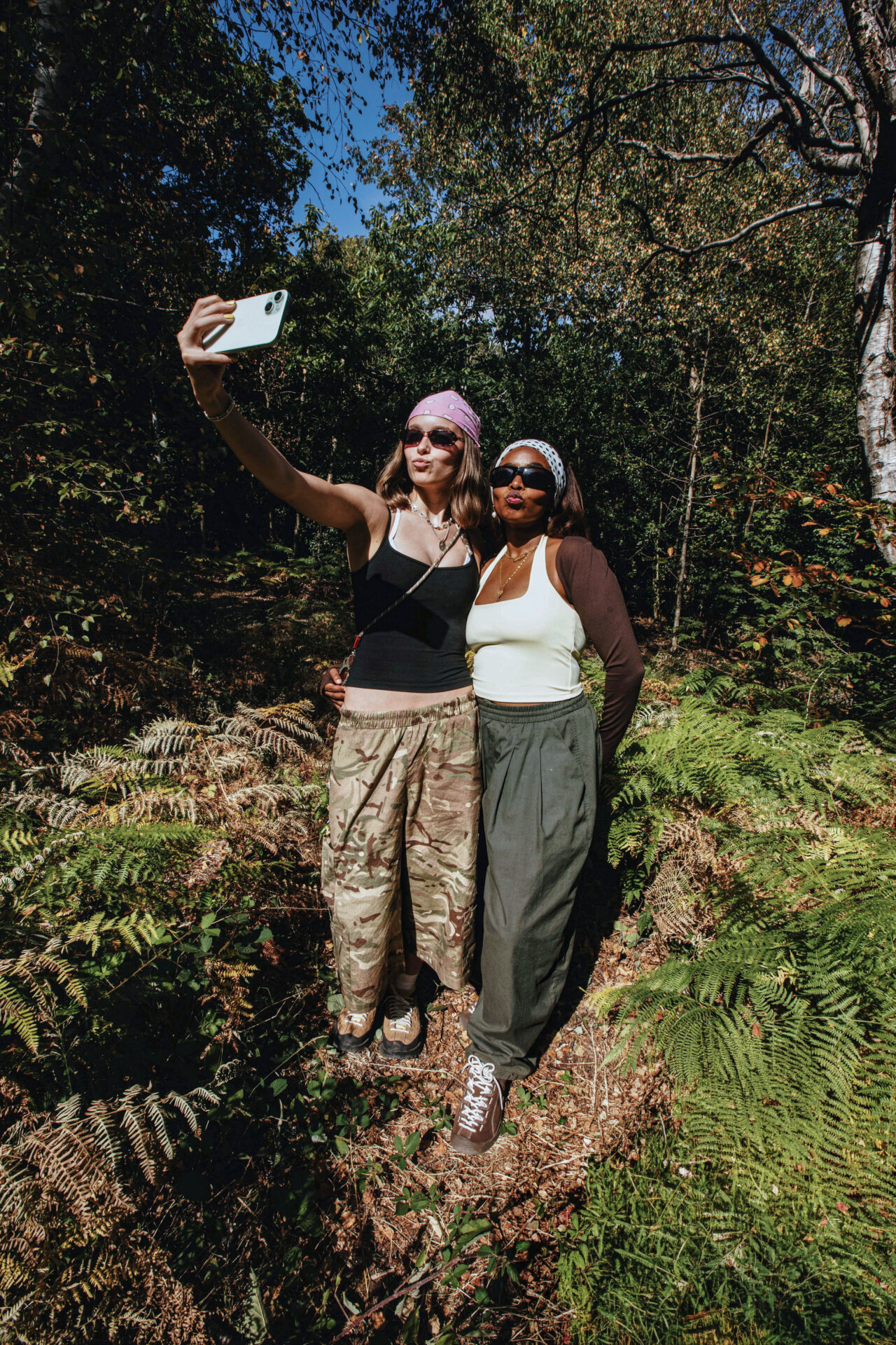 Two people posing for a selfie outside. One wears khaki parachute pants, a white tank top, brown bolero style top, brown KEEN Jaspers, and black sunglasses; the other wears the beige KEEN Jasper Zionic with white socks, a black tank top, camo print jorts, and black sunglasses.