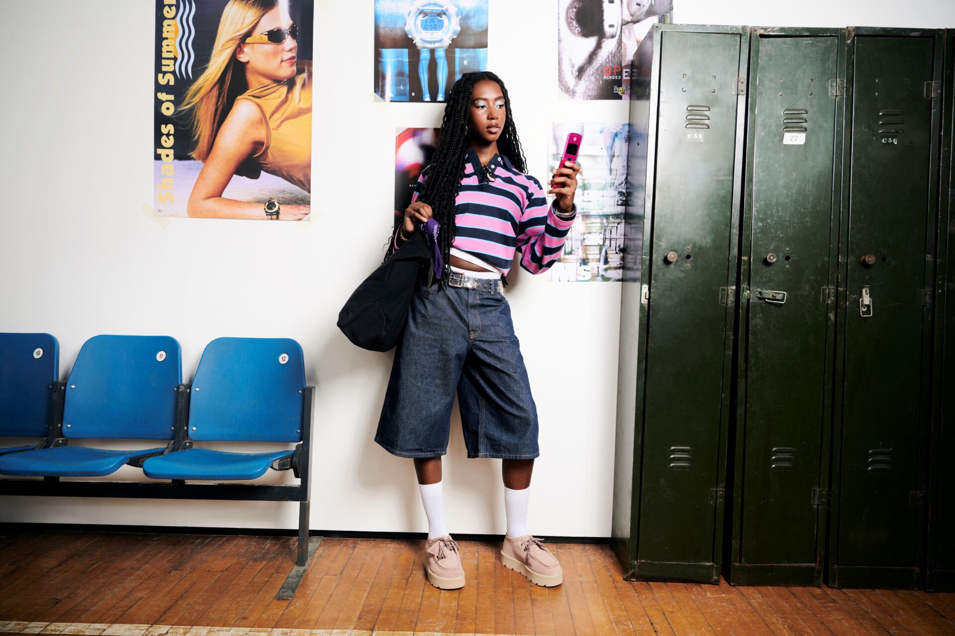 Person wearing blue jorts, a striped blue and pink polo long sleeve, white socks, and the pink Clarks Wallabee, holding a pink flip phone.